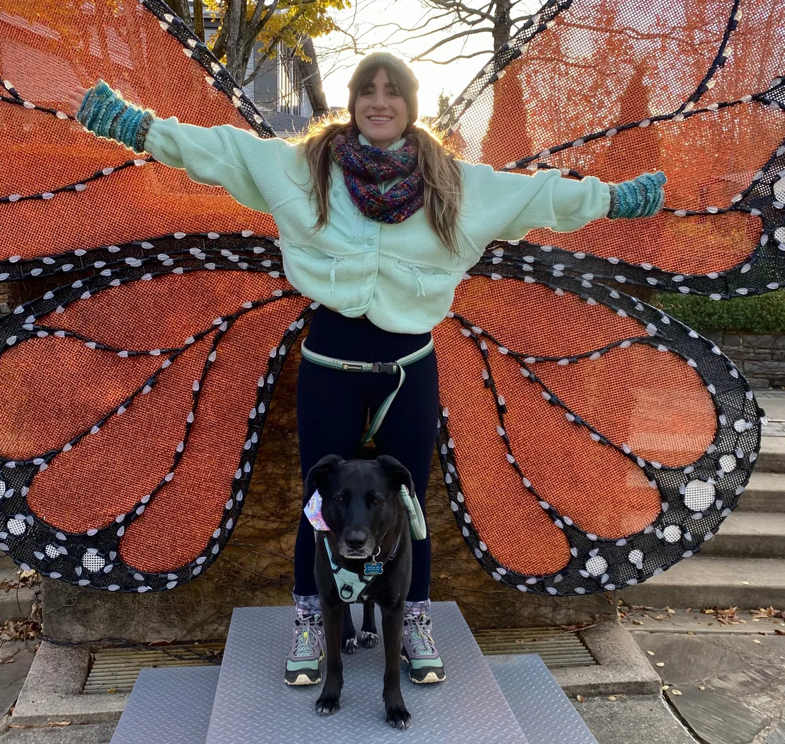 Lauren standing with her arms outstretched in front of a large monarch butterfly mural, her dog in front of her