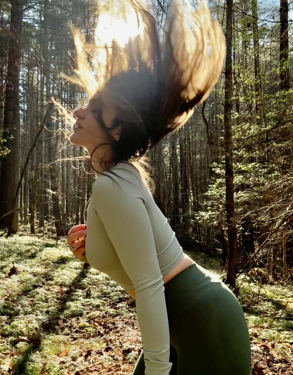 Lauren throwing her hair back in a sunlit forest, the sun catching it mid-flip