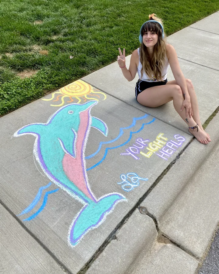 Lauren in headphones, sitting beside a large chalk drawing of a dolphin she made on the sidewalk